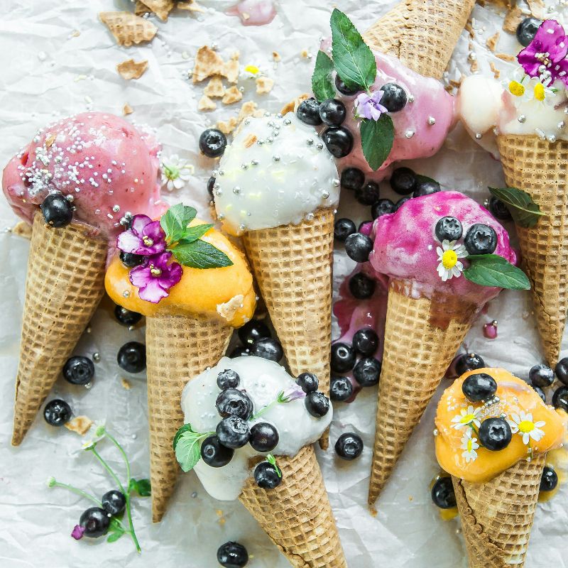 Cornets de crème glacée colorés garnis de petits fruits, fleurs comestibles et perles sucrées, parfaits pour l’été.