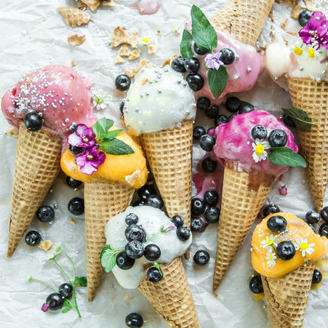 Cornets de crème glacée colorés garnis de petits fruits, fleurs comestibles et perles sucrées, parfaits pour l’été.